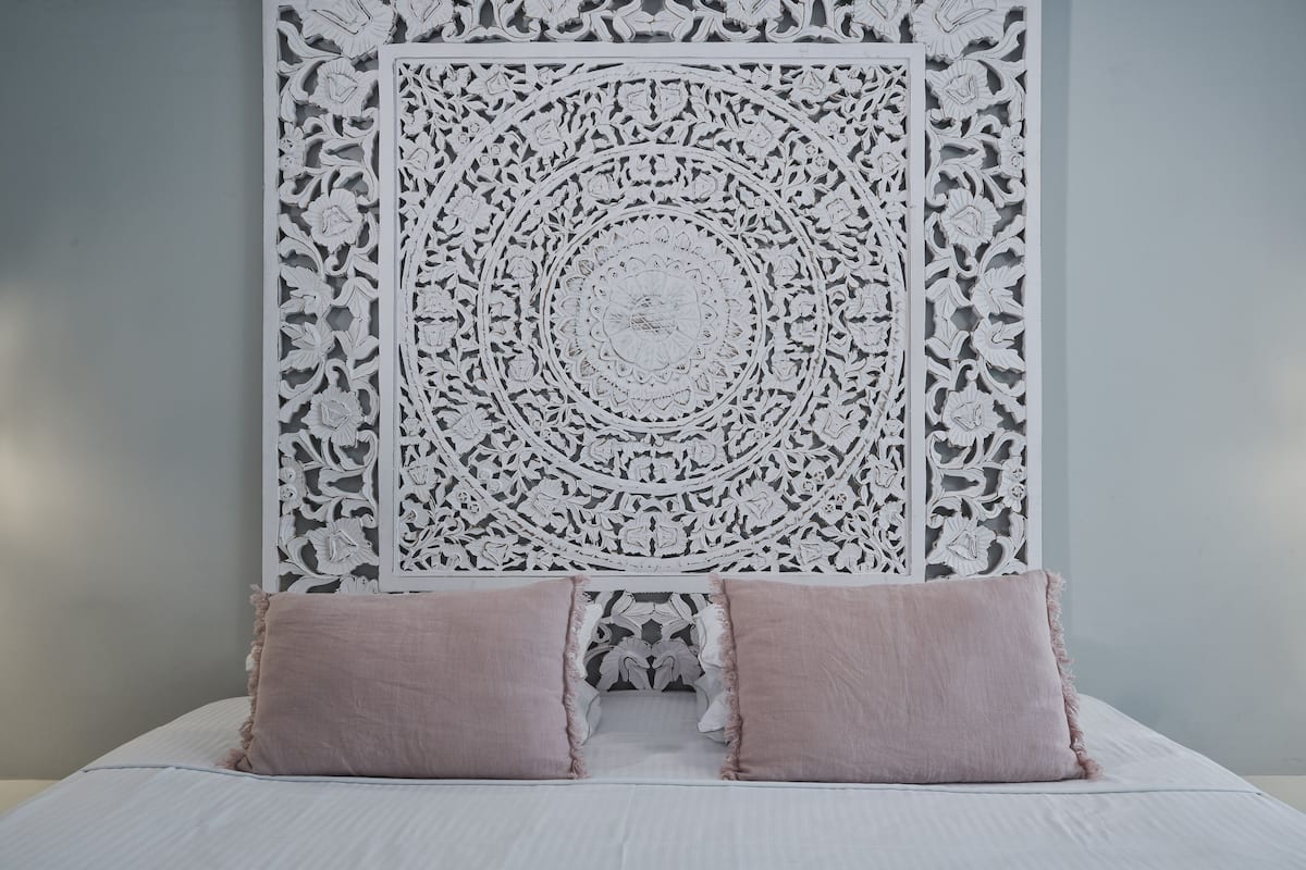 A bed with a white sheet and two pink pillows, a white decorative wall panel, and a grey wall.