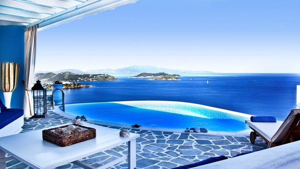 Une terrasse avec une piscine donnant sur une mer d’un bleu limpide, un sol en pierre et des chaises longues.
