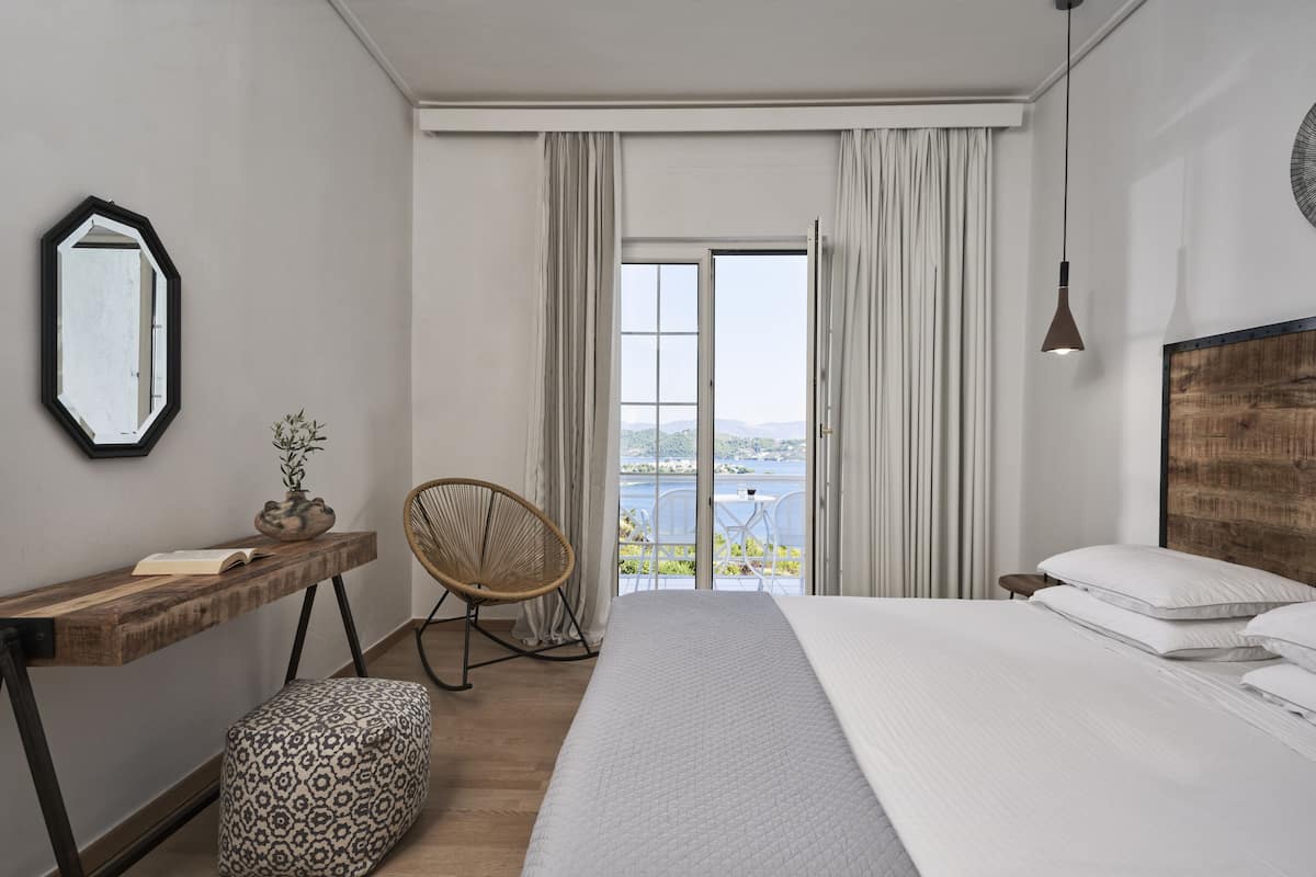 A bedroom with a large bed, a wooden desk, a chair, and a view of a balcony with a table and chairs.
