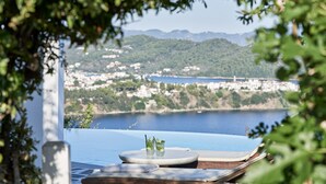 Un espace aménagé au bord de la piscine, avec vue sur une ville côtière, une table avec des boissons et des chaises longues.