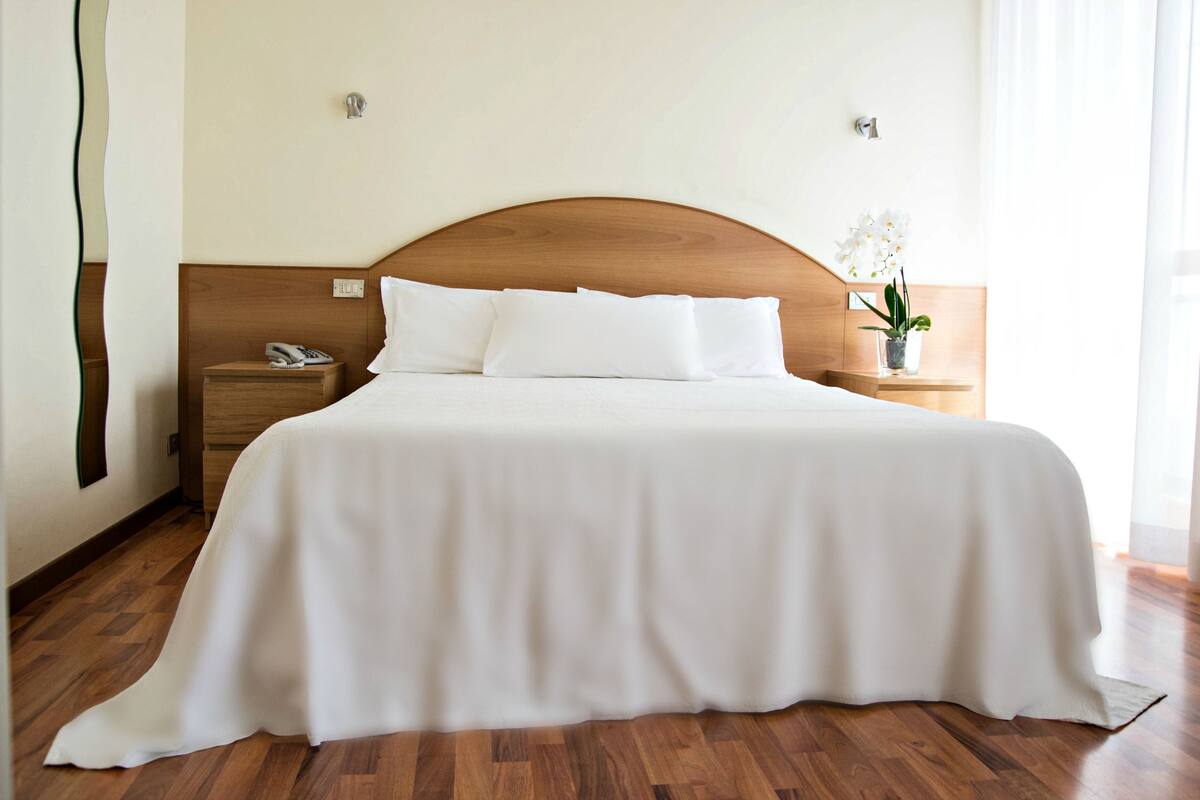 Ein ordentlich bezogenes Bett in einem Hotelzimmer, mit einem hölzernen Kopfteil, einem Nachttisch mit Telefon und einem kleinen Topfpflanzen.