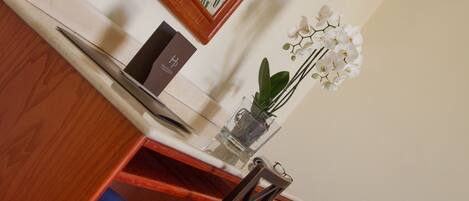 A hotel room with a wooden desk, a mirror, a vase with flowers, a telephone, and a small bedside table.