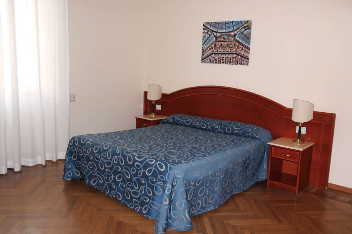 Ein Schlafzimmer mit einem Holzkopfende, einer blau gemusterten Bettdecke, zwei Nachttischen mit Lampen und einem gerahmten Bild an der Wand.