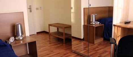 Une chambre d’hôtel avec un plancher en bois, un lit recouvert d’une couverture bleue, une table de chevet avec une lampe, un petit tabouret en bois et un miroir accroché au mur.