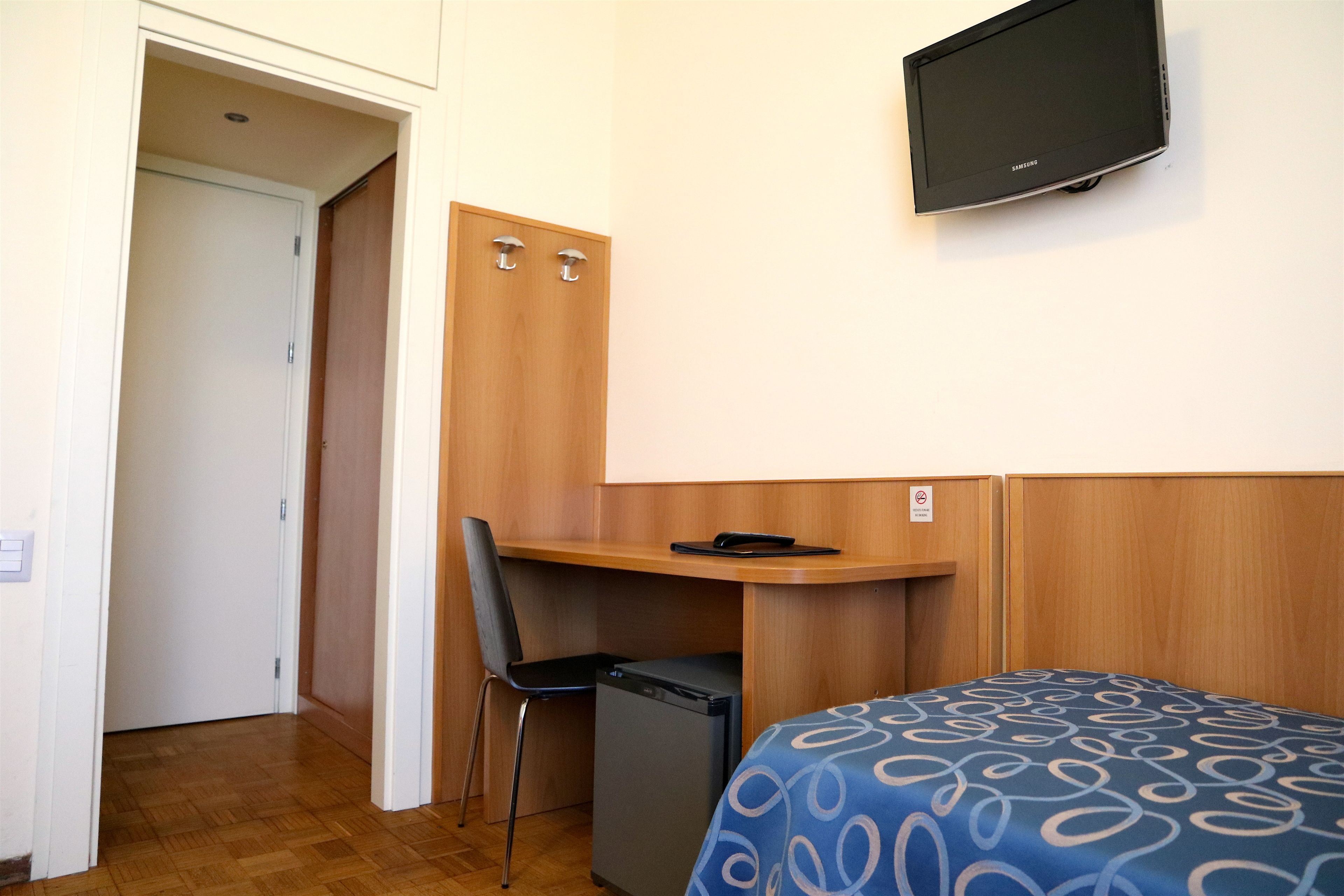 A hotel room with a bed, a desk, a chair, a small refrigerator, and a television mounted on the wall.