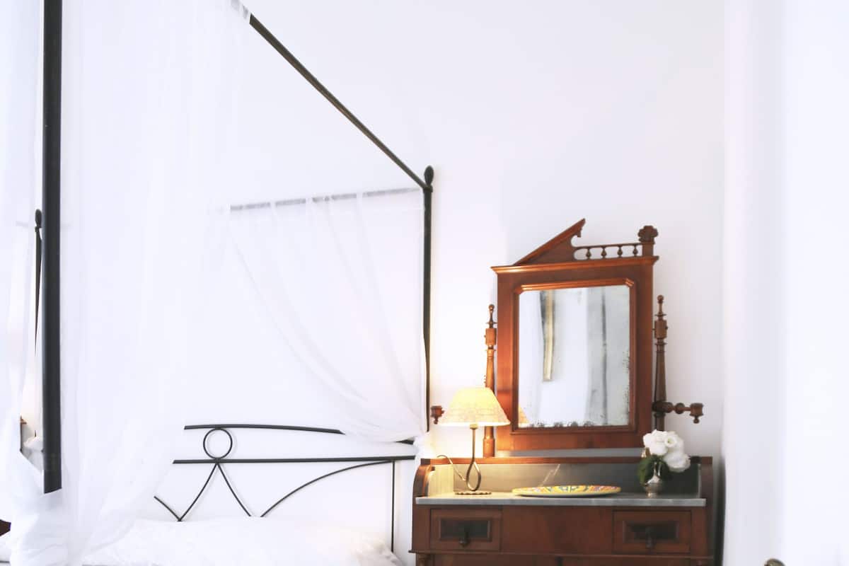 Ein Zimmer mit einem Bett, einem Nachttisch mit Lampe, einem Spiegel, einem blumigen Sessel und einem kleinen Tisch mit Tassen und einem Löffel.
