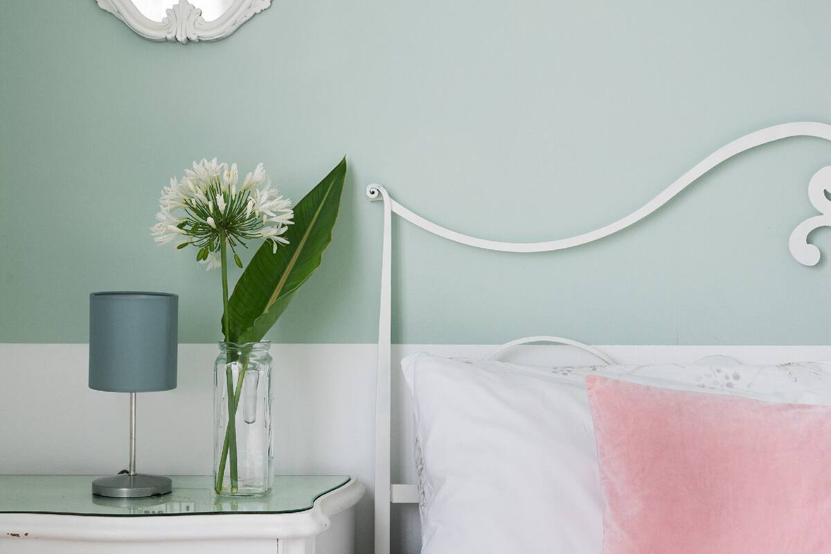 Ein Schlafzimmer mit einem weißen Bett, einem weißen Nachttisch, einem Spiegel, einer Lampe und einer Vase mit Blumen.