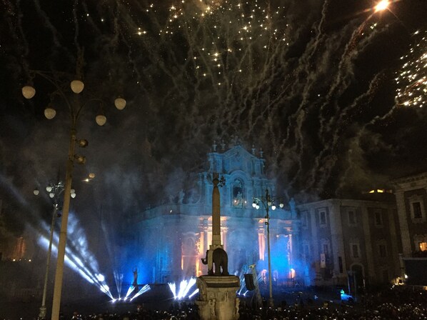 A nighttime view of a fountain with water jets and a statue, illuminated by blue and white lights, with a building in the background.