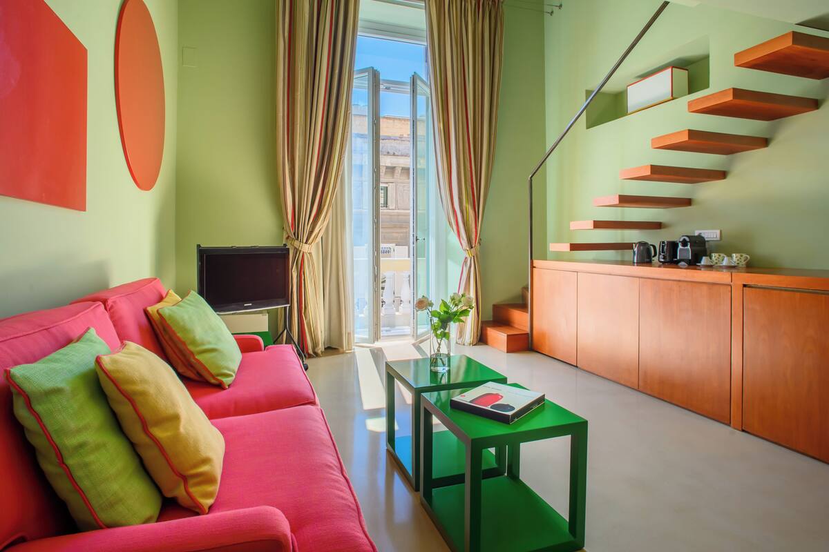 Una sala de estar moderna con un sofá rojo, una mesa de centro verde y una televisión. Hay cortinas, una escalera y vistas al exterior.