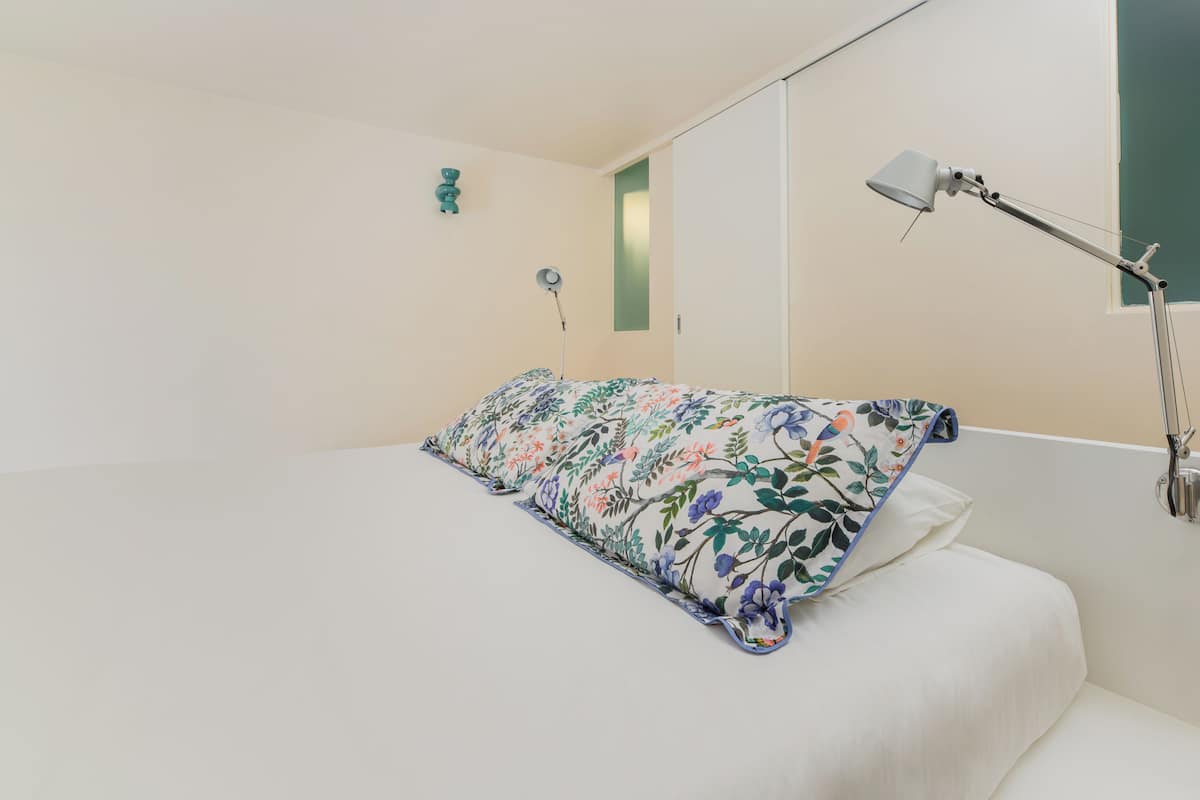 Una cama bien hecha con almohadas estampadas con motivos florales, una lámpara de noche y un pequeño adorno en la pared.