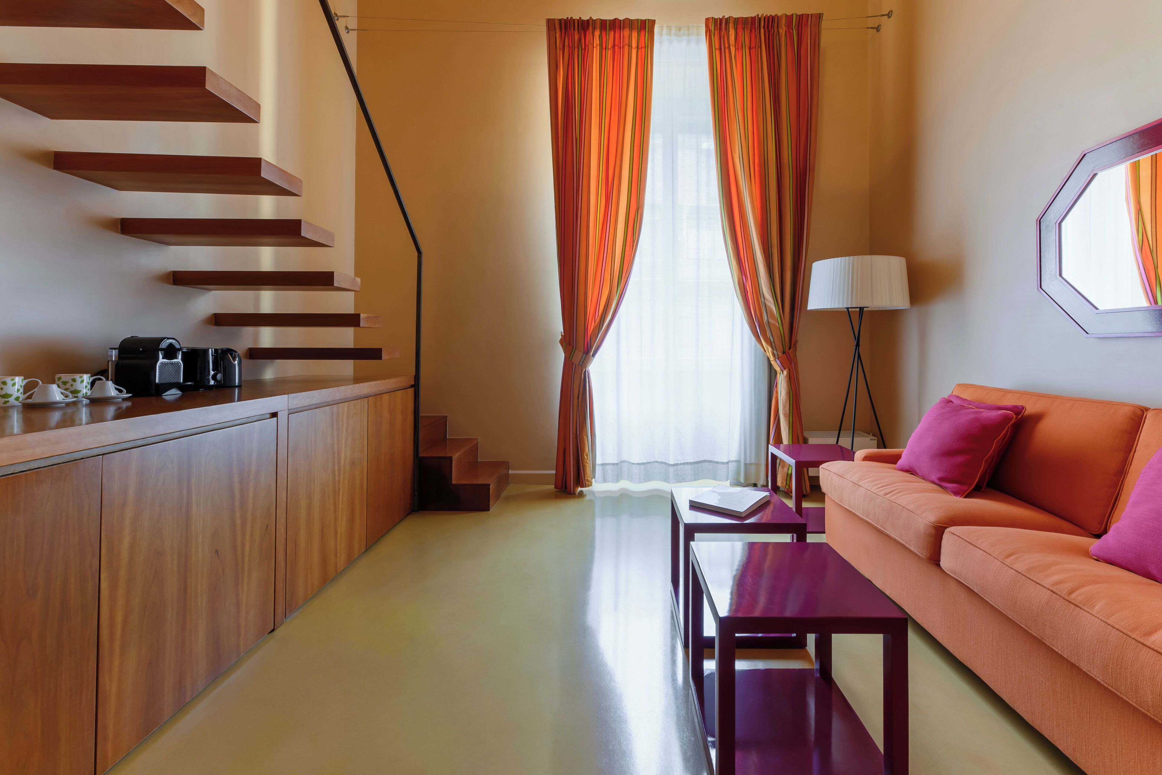 A modern hotel room with a wooden staircase, a sofa, a coffee table, and a desk with a lamp.