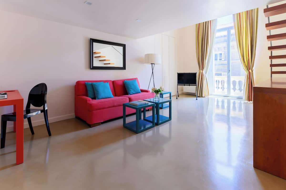 Una sala de estar moderna con un sofá rojo, dos mesitas auxiliares azules, un sillón negro, un cuadro en la pared y un ventanal con cortinas.