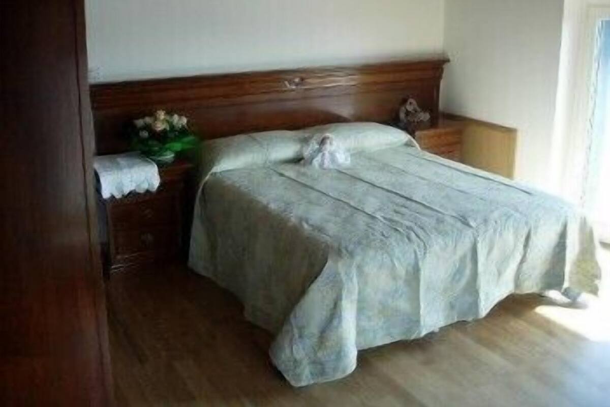 A bedroom with a wooden headboard, a single bed with a light-colored bedspread, a nightstand with a vase of flowers, and a wooden dresser.