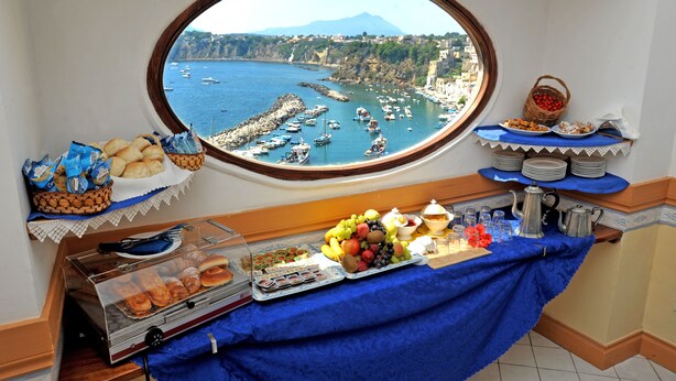 A breakfast buffet with a scenic coastal view through a window, including boats and a mountain in the background. - La Casa sul Mare (Procida)