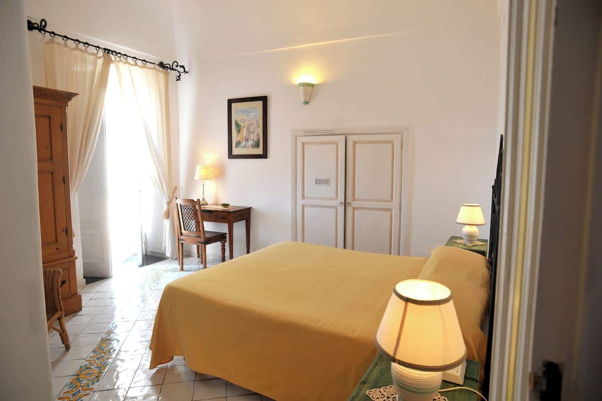 A hotel room with a bed, two bedside tables with lamps, a chair, a desk with a chair, a wardrobe, and a door.