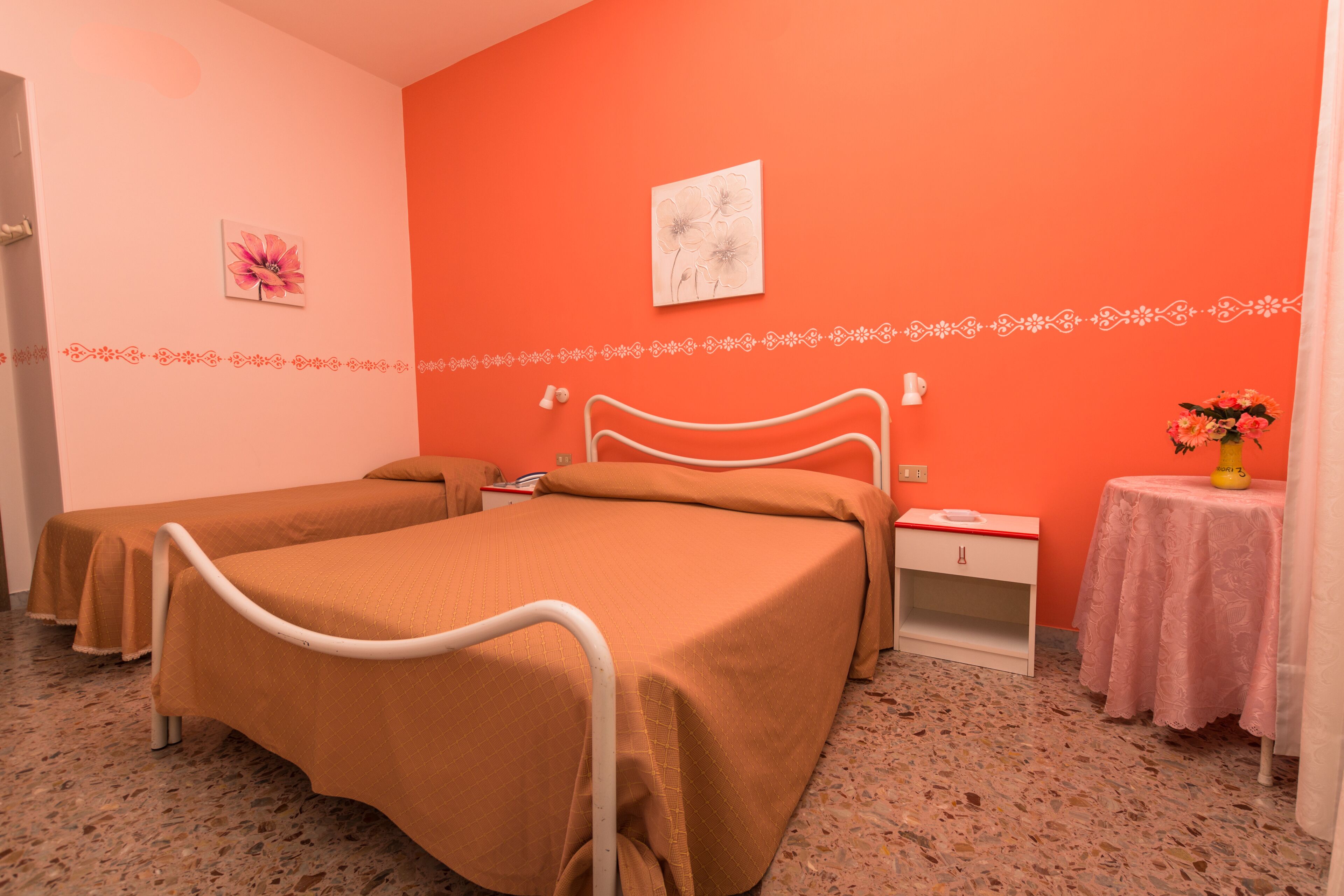 Habitación de hotel con dos camas, una mesita con un florero y una pared decorada con un cuadro floral.