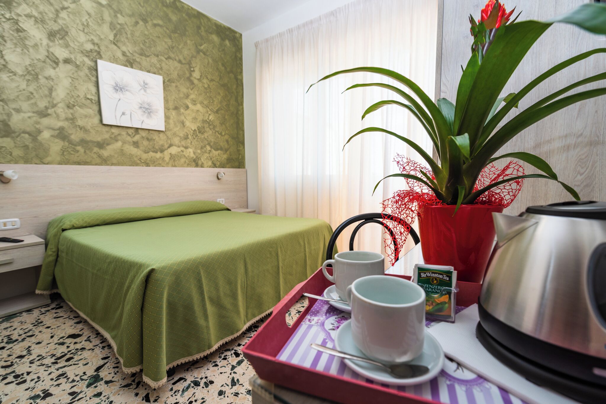 A hotel room with a bed, a tray with cups and a kettle, a plant, and a painting on the wall.