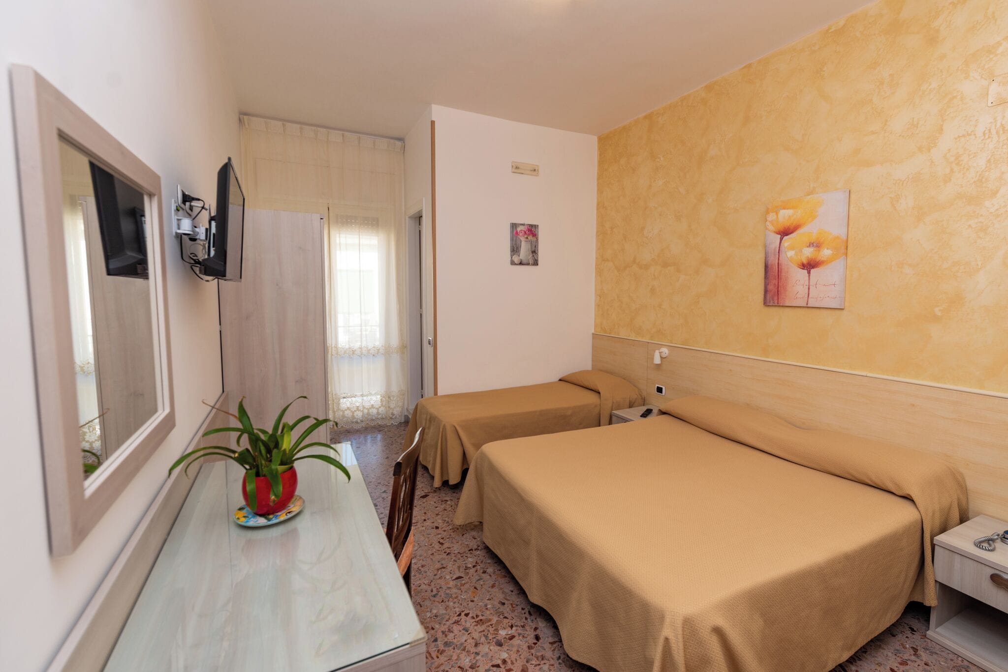 A hotel room with two single beds, a small table with a potted plant, a TV mounted on the wall, and a painting of flowers on the wall.