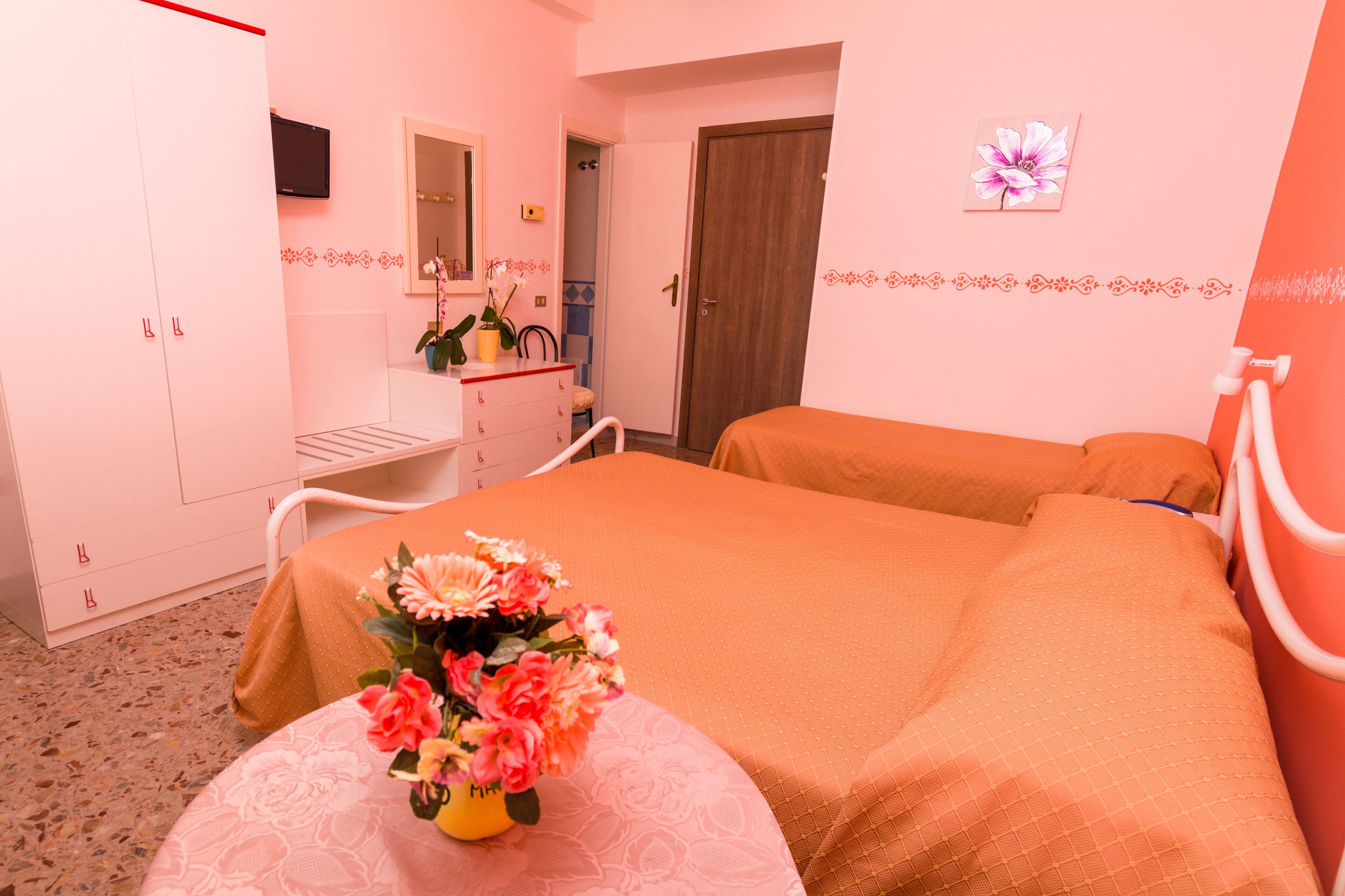 A hotel room with a bed, a bedside table with a vase of flowers, a wardrobe, a mirror, and a door.