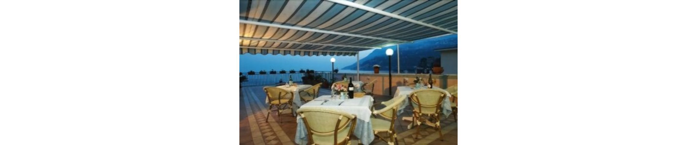 An outdoor dining area with tables and chairs, covered by a striped awning, and a view of the sea in the background.