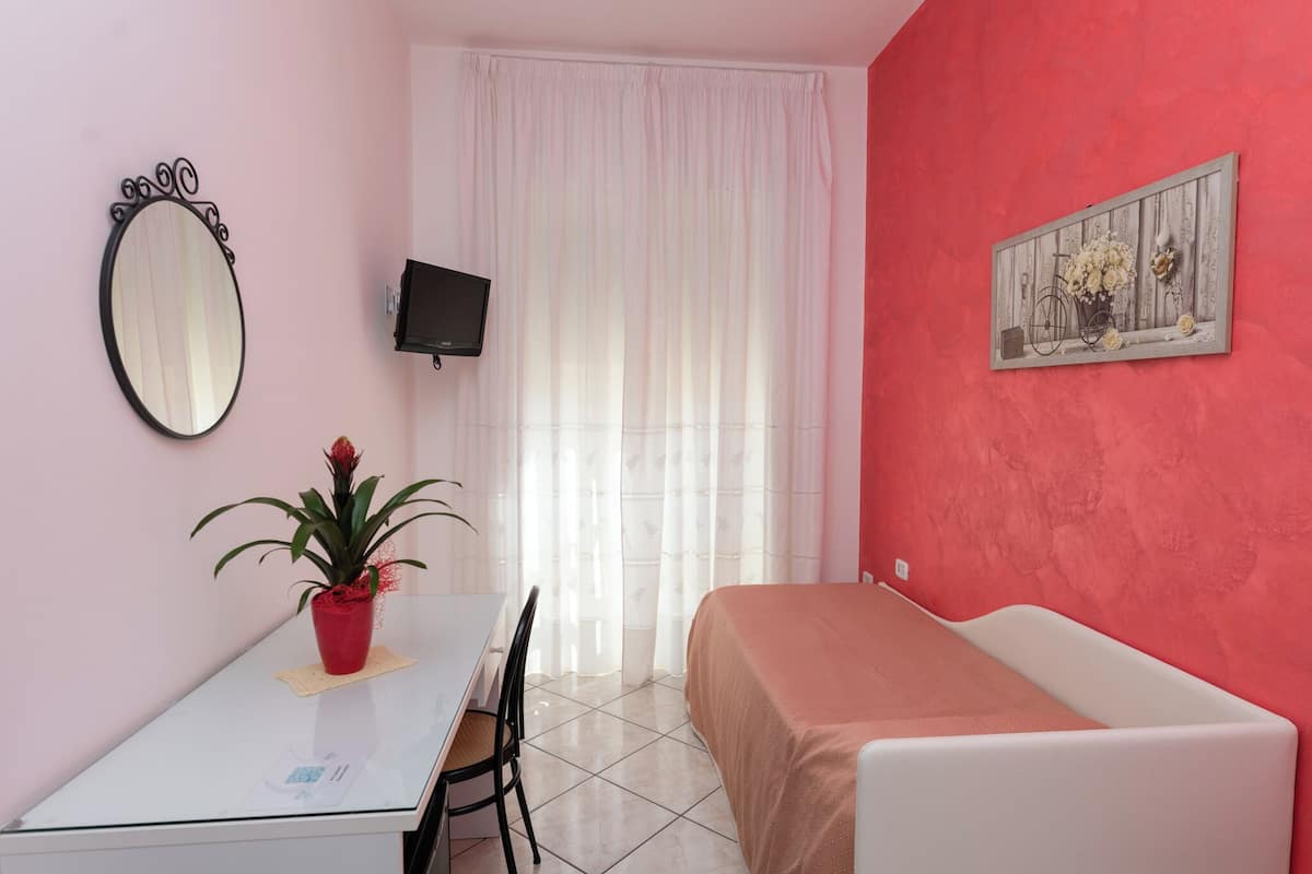 A small, tidy room with a bed, a table with a chair, a potted plant, a television, and a window with curtains.