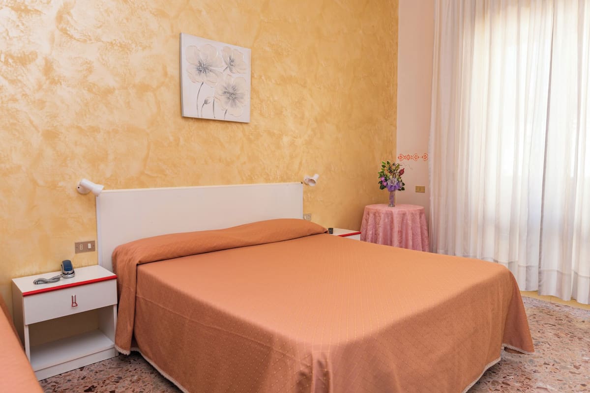 A hotel room with a bed, a bedside table with a lamp, a small table with a vase of flowers, and a window with curtains.