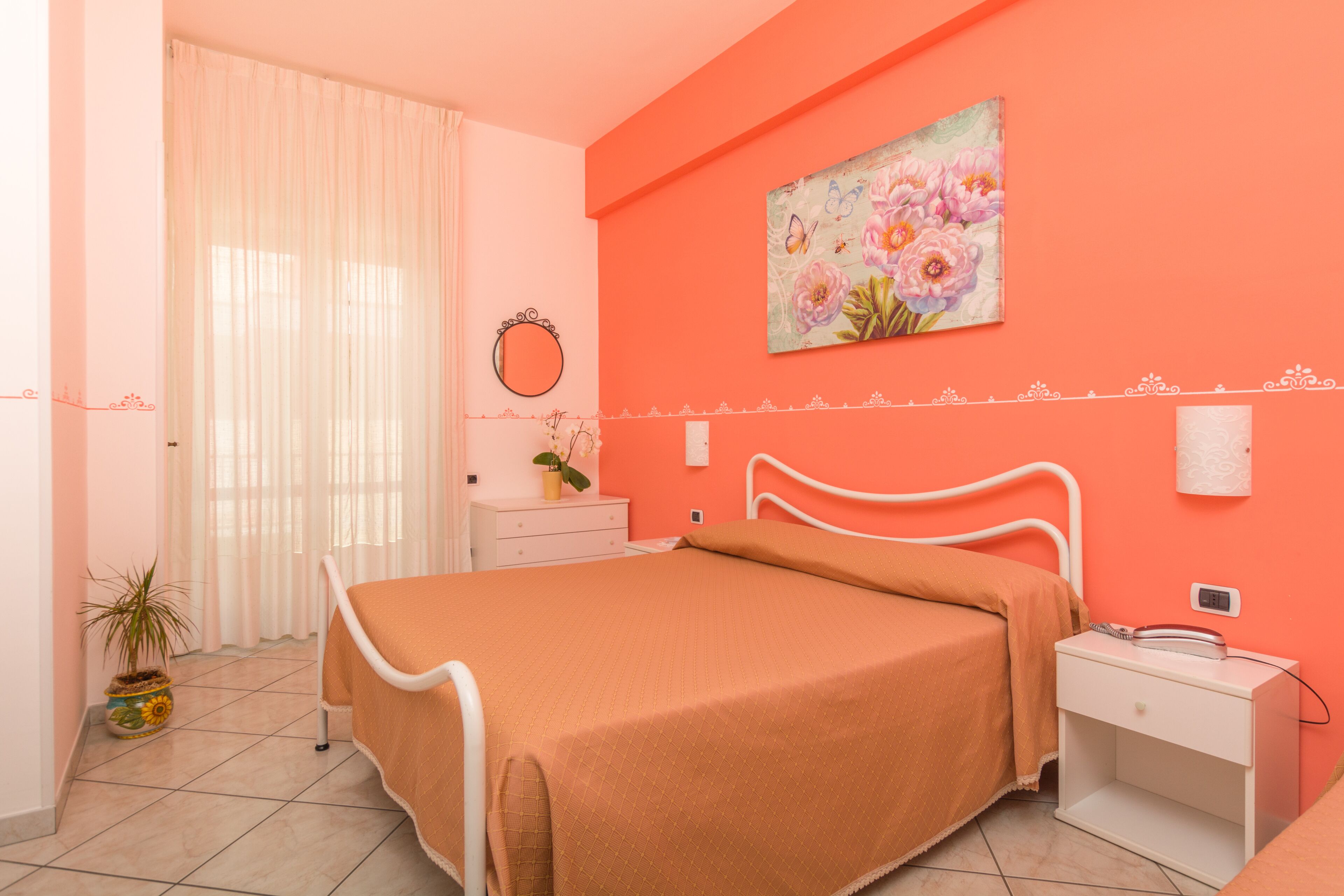 A bedroom with a bed, a nightstand, a bedside table, a potted plant, a mirror, and a floral painting on the wall.