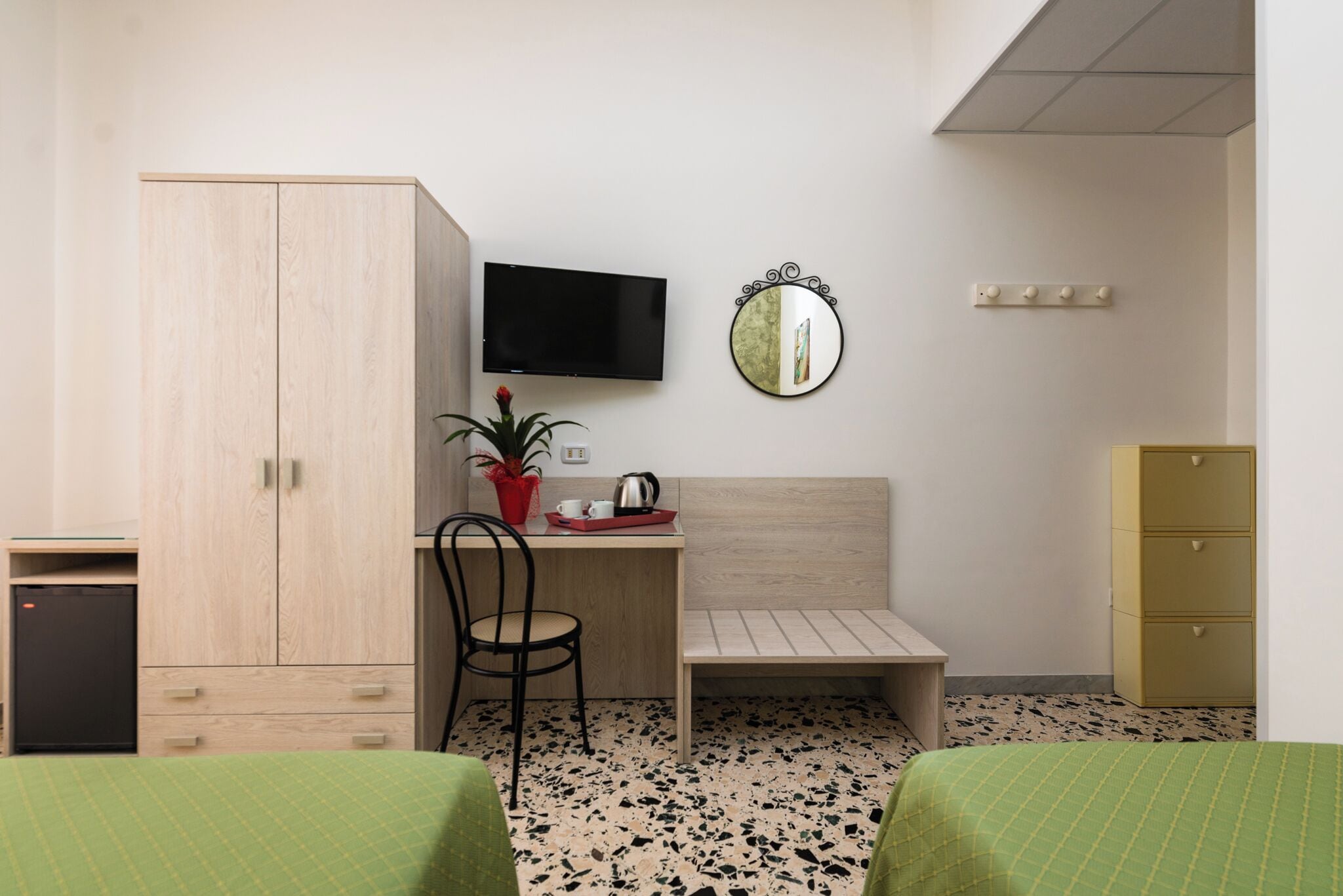A bedroom with a bed, a desk, a chair, a wardrobe, a television, a mirror, and a small cabinet.