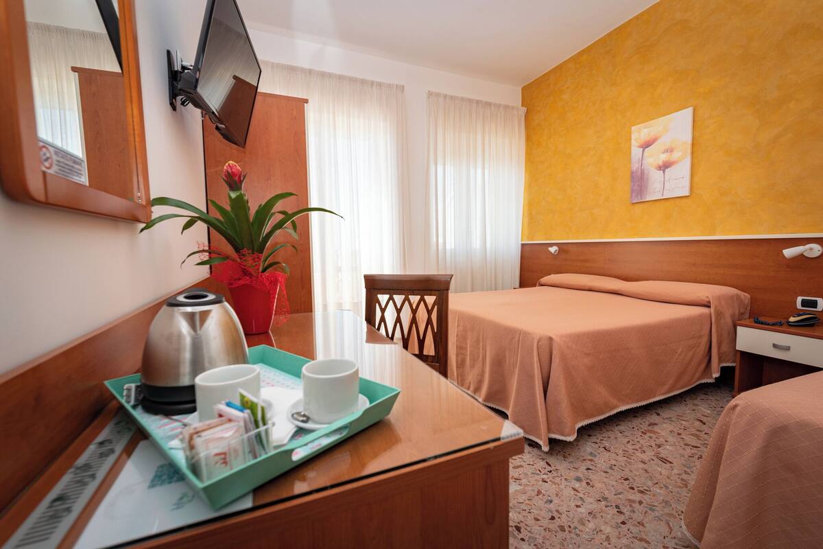 A hotel room with a bed, a desk with a tray of breakfast items, a potted plant, and a painting on the wall.