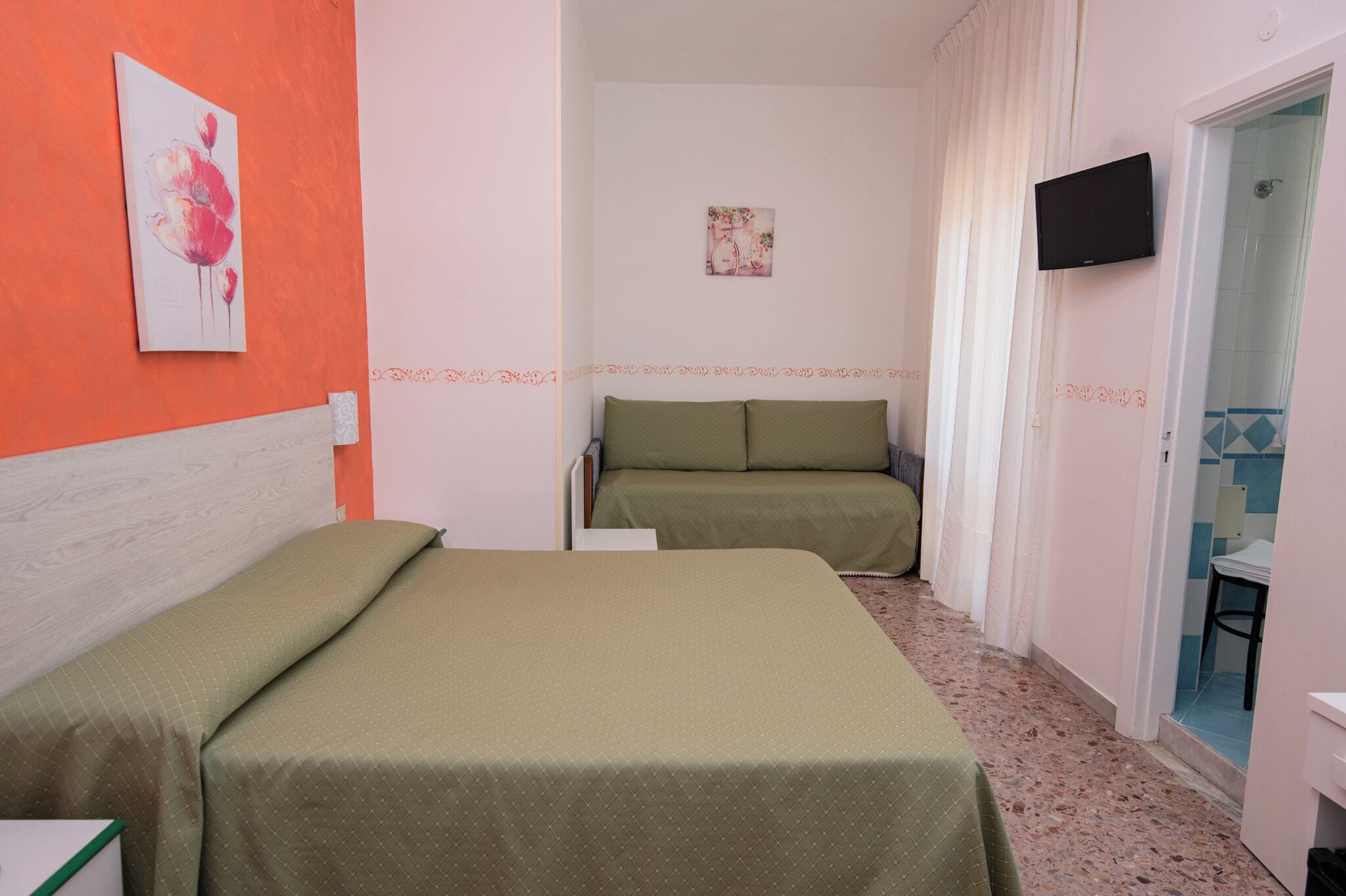 A hotel room with a bed, a sofa, a TV mounted on the wall, and a bathroom visible through an open door.
