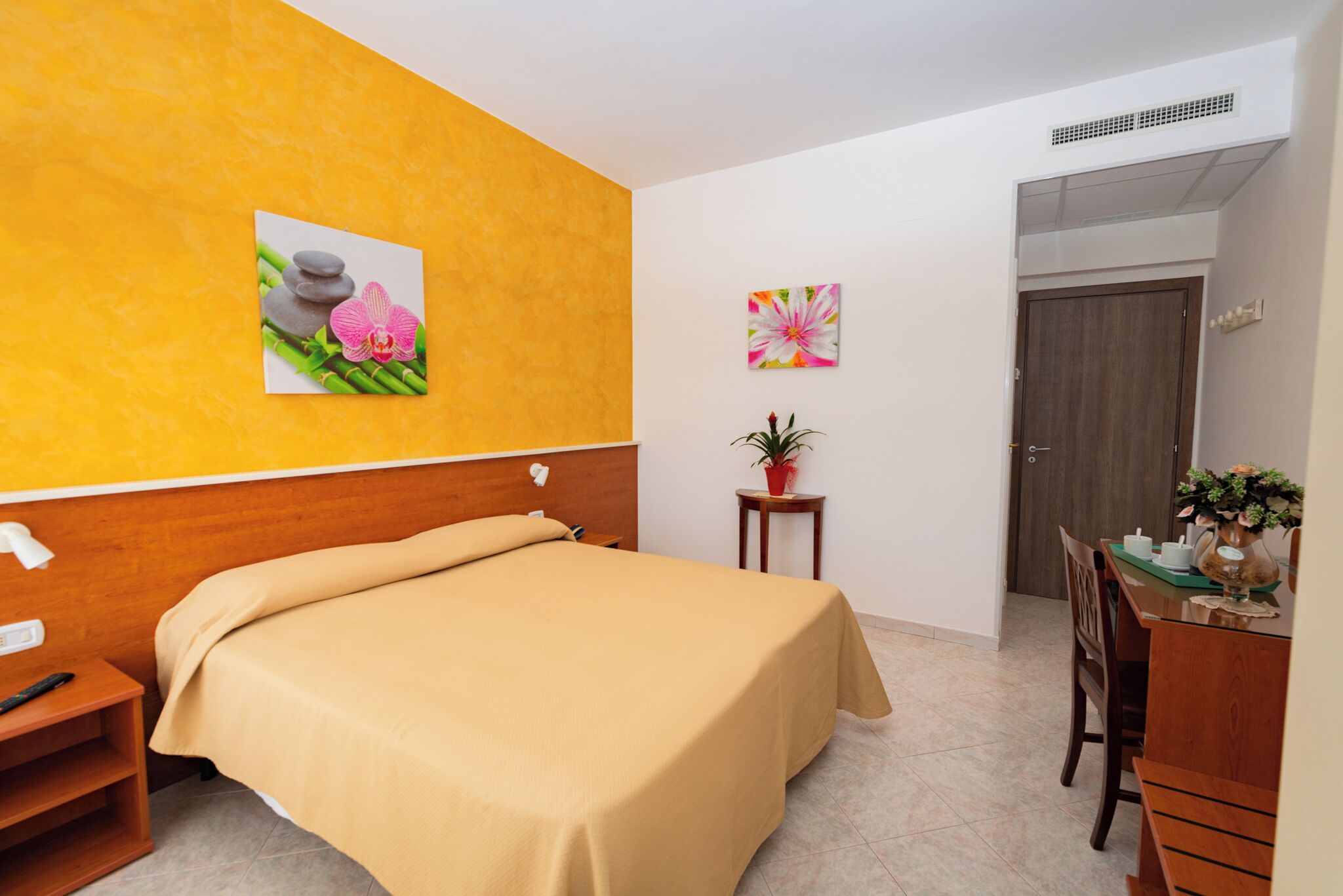 Habitación de hotel con una cama, una mesita con un florero y una planta en maceta, y una puerta que da a otra habitación.