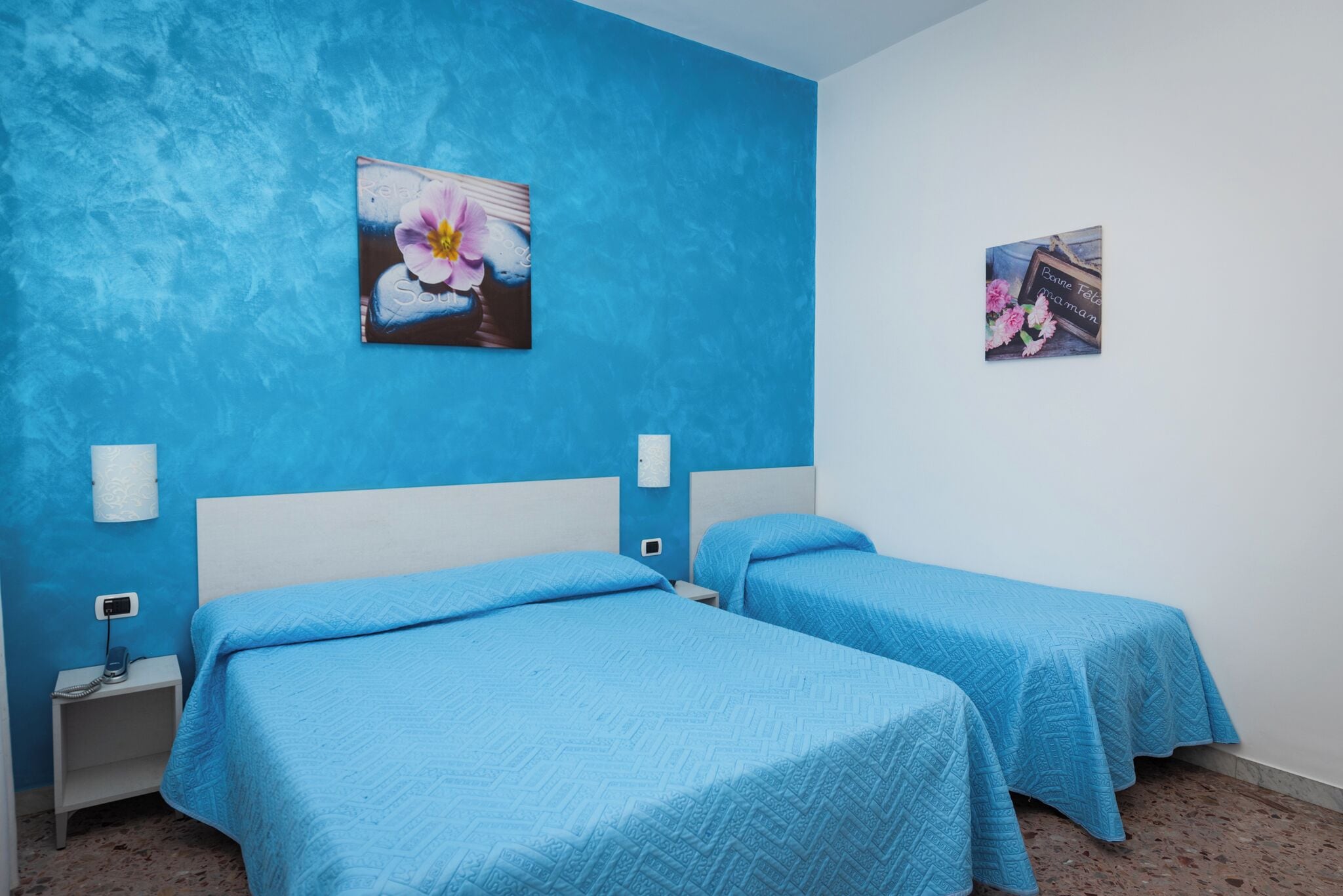 A room with two beds, each with a blue bedspread and headboard, a nightstand with a phone, and a wall with two floral paintings.