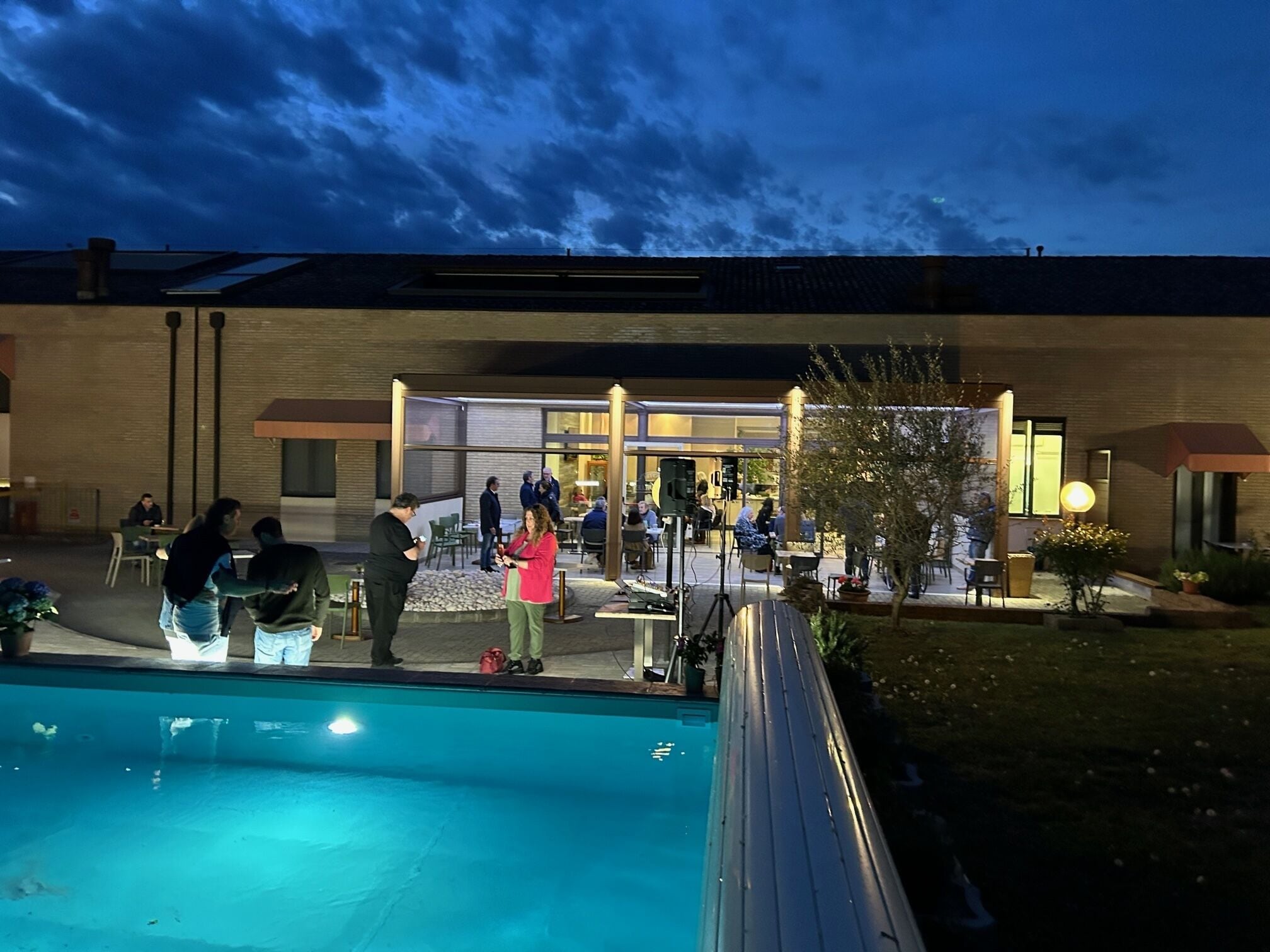 Area piscina all'aperto con persone riunite, un edificio con ingresso in vetro e un patio ben illuminato.