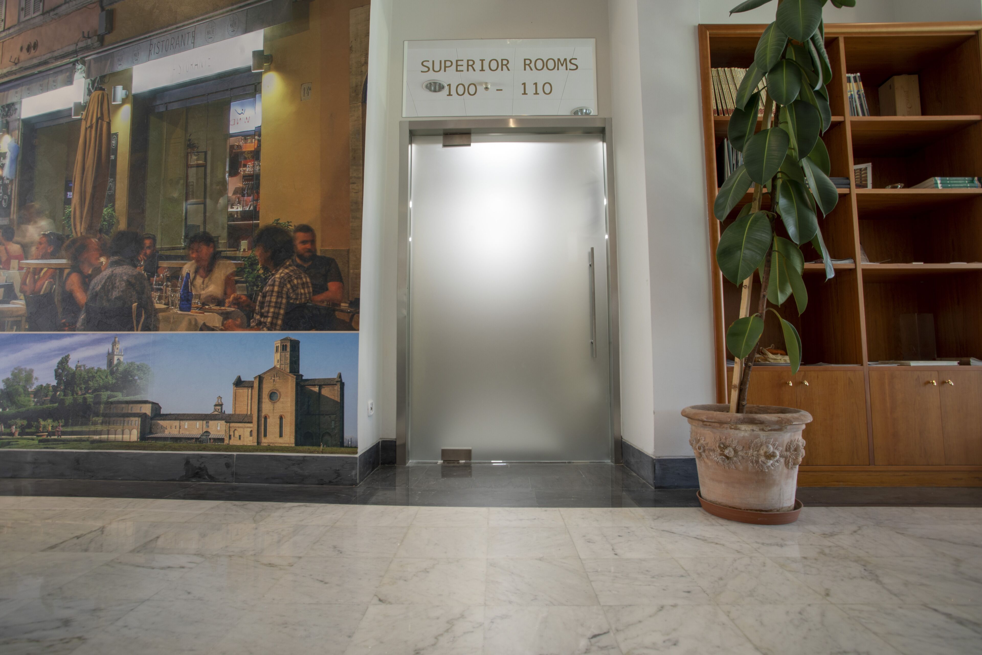 En hotelllobby med en skylt som visar vägen till ”rumskategorin Superior, rum 100–110”, en krukväxt, en bokhylla och en väggmålning som föreställer en byggnad.