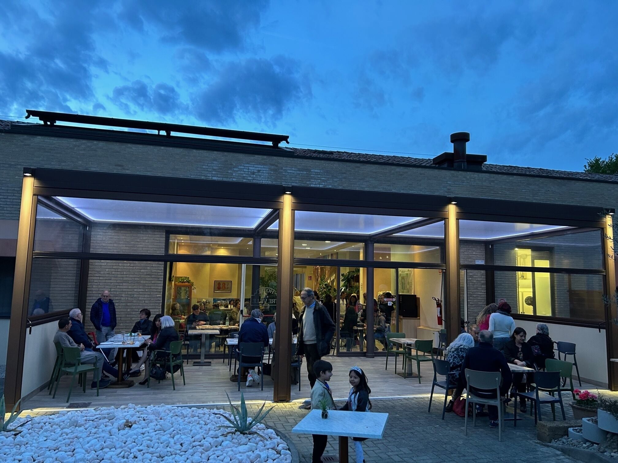 Un café al aire libre con mesas y sillas, un cerramiento de vidrio y un edificio de ladrillo al fondo.