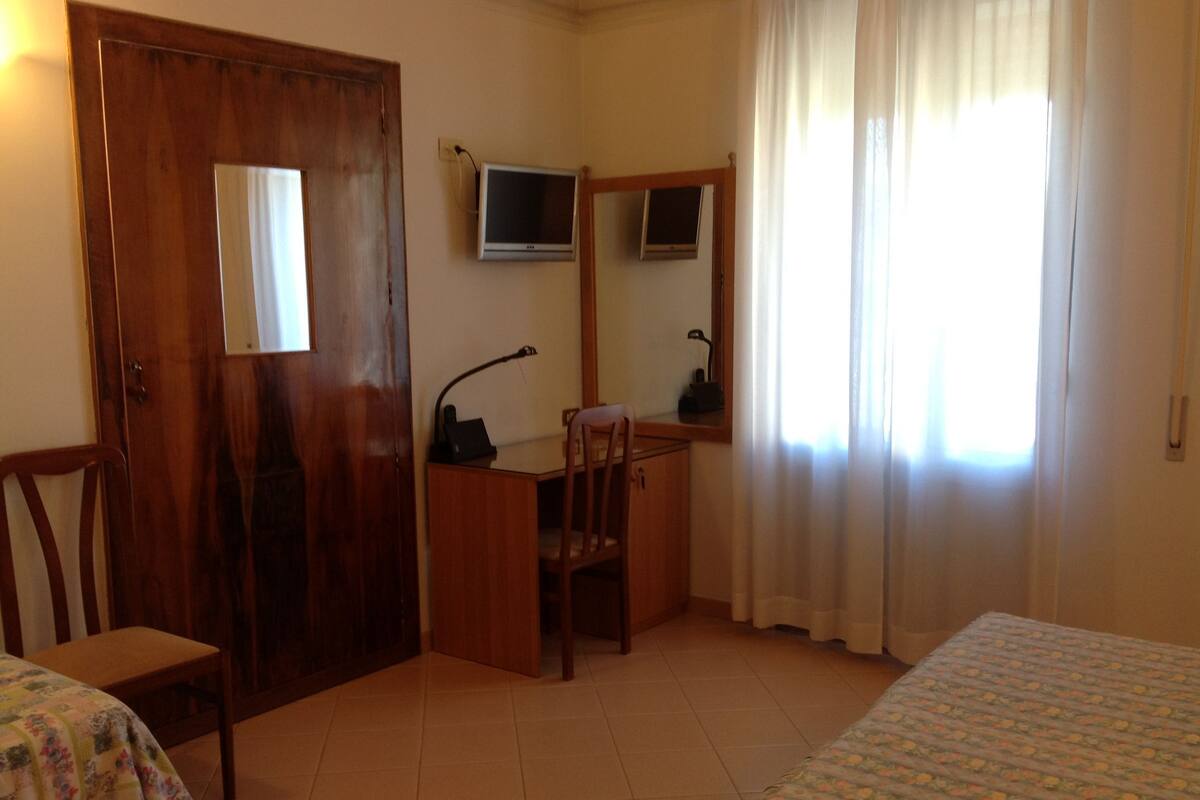 Una habitación con escritorio de madera, una silla, una cama, un televisor y un ventanal con cortinas.