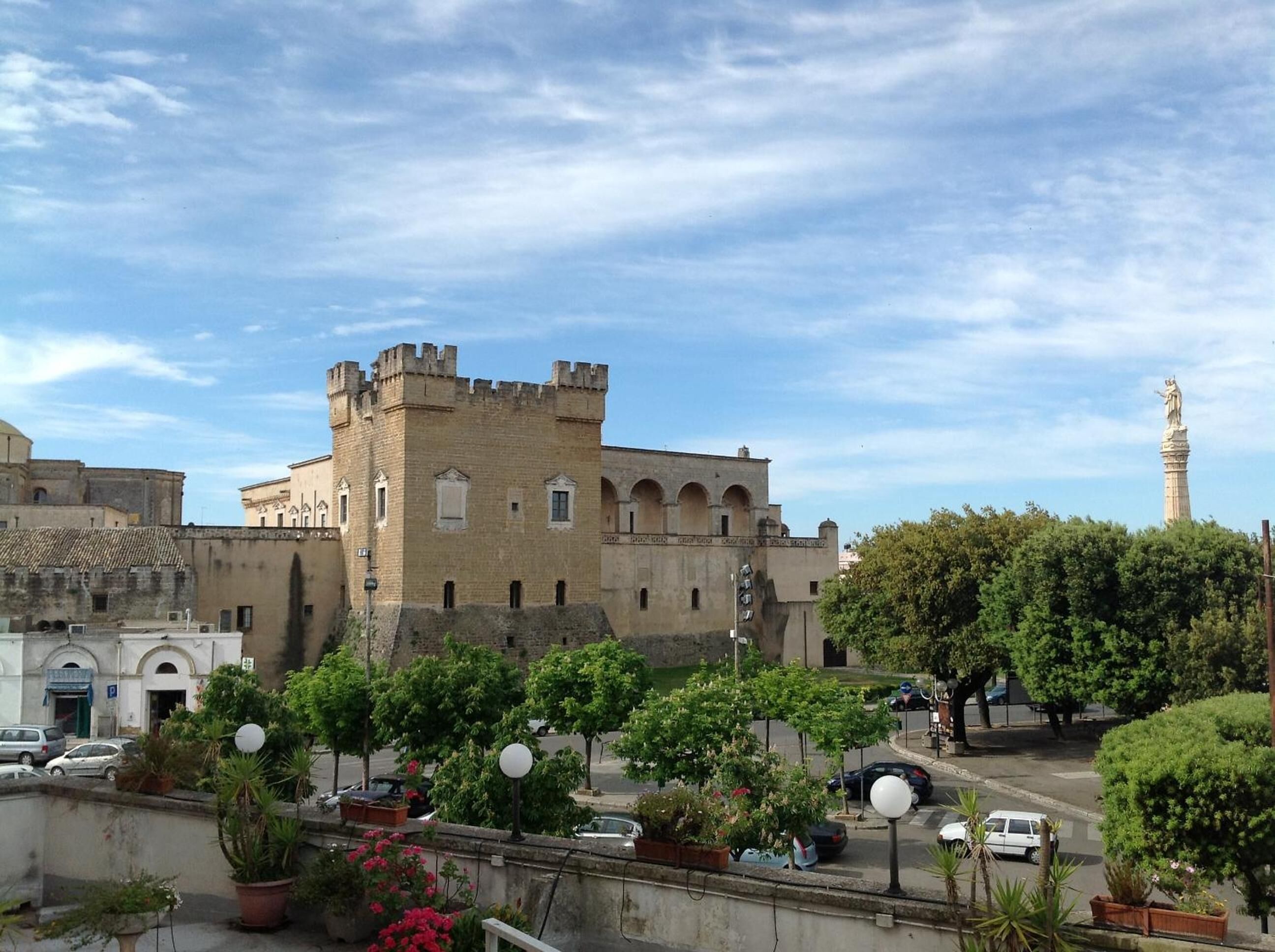 Un edificio histórico con una torre destacada y una estatua, rodeado de vegetación y coches aparcados.