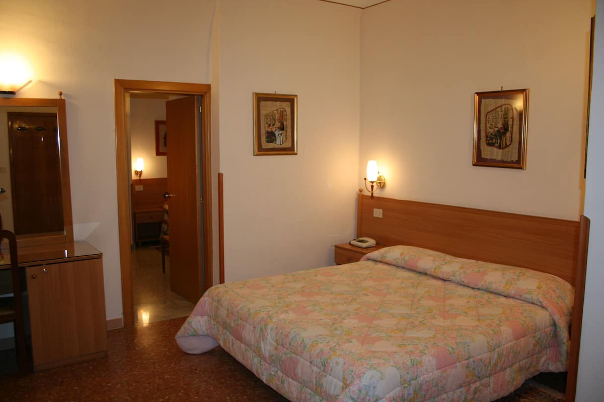 Un dormitorio con cama de madera, una mesita de noche, una lámpara de noche, un armario y una puerta que da a otra habitación.