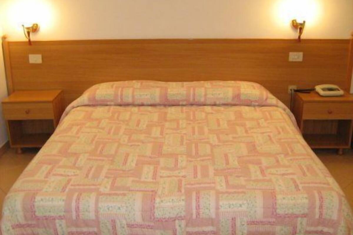 Habitación estándar de hotel con cabecera de madera, colcha estampada, dos mesitas de noche con lámparas y un teléfono sobre una de ellas.