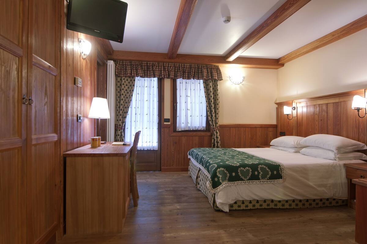 Una habitación con paneles de madera, una cama, un escritorio, una silla, un televisor y dos ventanas con cortinas.