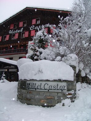 A snow-covered Hotel Castor sign with a stone base, surrounded by snow-laden trees and a building with red-framed windows in the background. - Hotel Castor (Ayas)