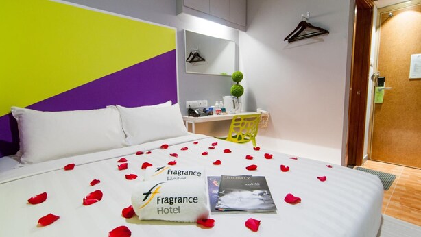 A hotel room with a bed covered in rose petals, a white pillow, and a towel with the hotel's logo. There is a small table with a coffee maker and a green chair. - Fragrance Hotel - Rose (Singapore)