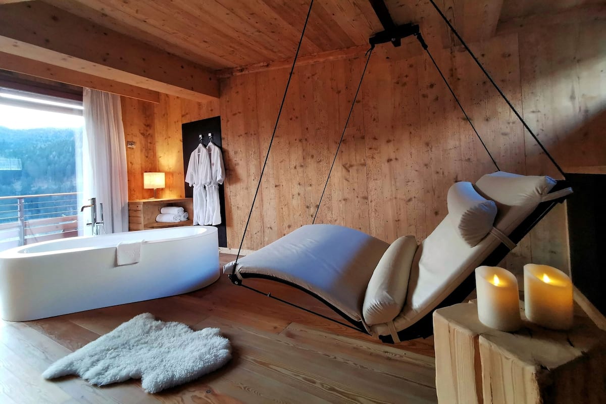 L’intérieur d’un chalet en bois, avec un fauteuil suspendu, une baignoire et une vue sur l’extérieur.