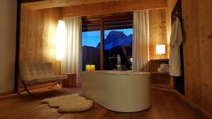 A wooden cabin interior with a central sink, a cushioned chair, a fur rug, and a mountain view through large windows.