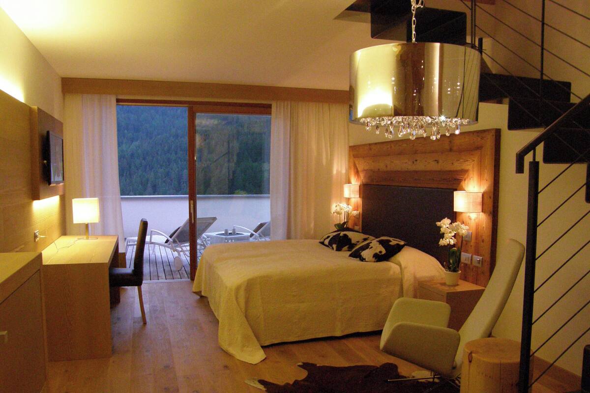 Une chambre d’hôtel comprenant un lit, un bureau avec une lampe, une chaise, une grande fenêtre avec des rideaux et un lustre.