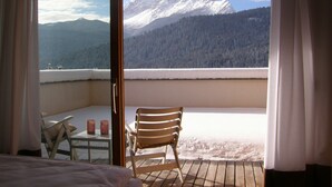 A balcony with a wooden chair and a small table, overlooking a snow-covered mountain and forest.
