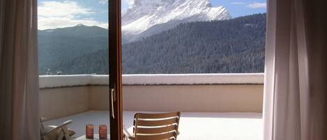 Un balcon avec une chaise en bois et une petite table, d’où l’on peut admirer une montagne et une forêt recouvertes de neige.