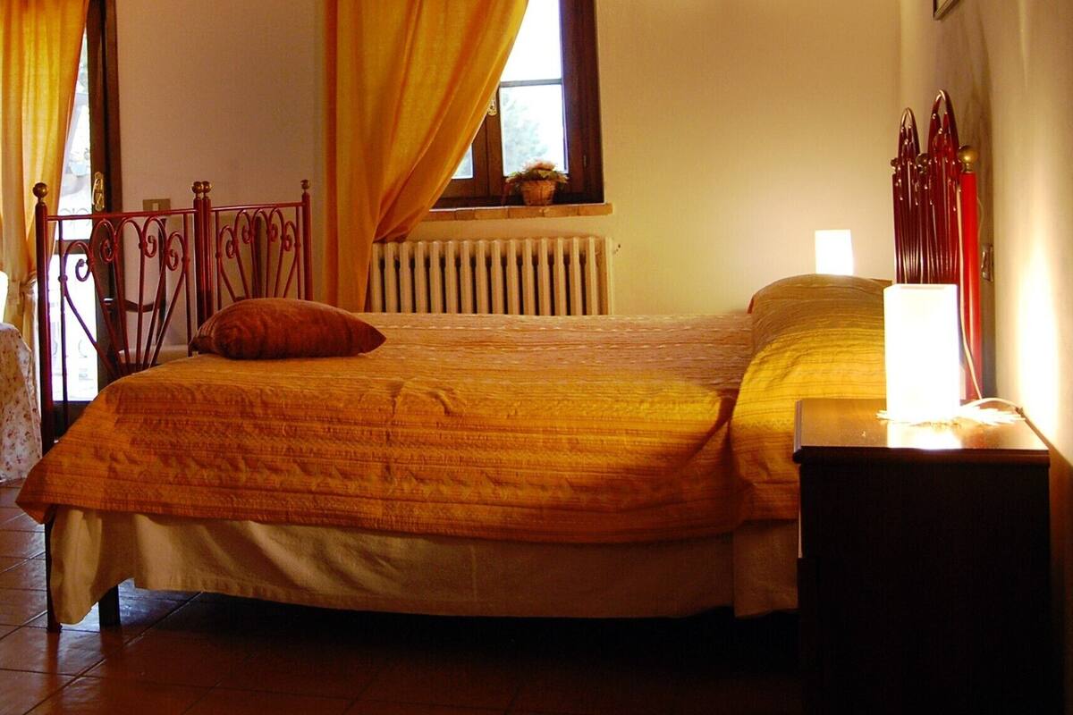 Une chambre avec un plafond en bois, un lit recouvert d’une couvre-lit à motifs, une table de chevet avec une lampe et une fenêtre avec des rideaux.