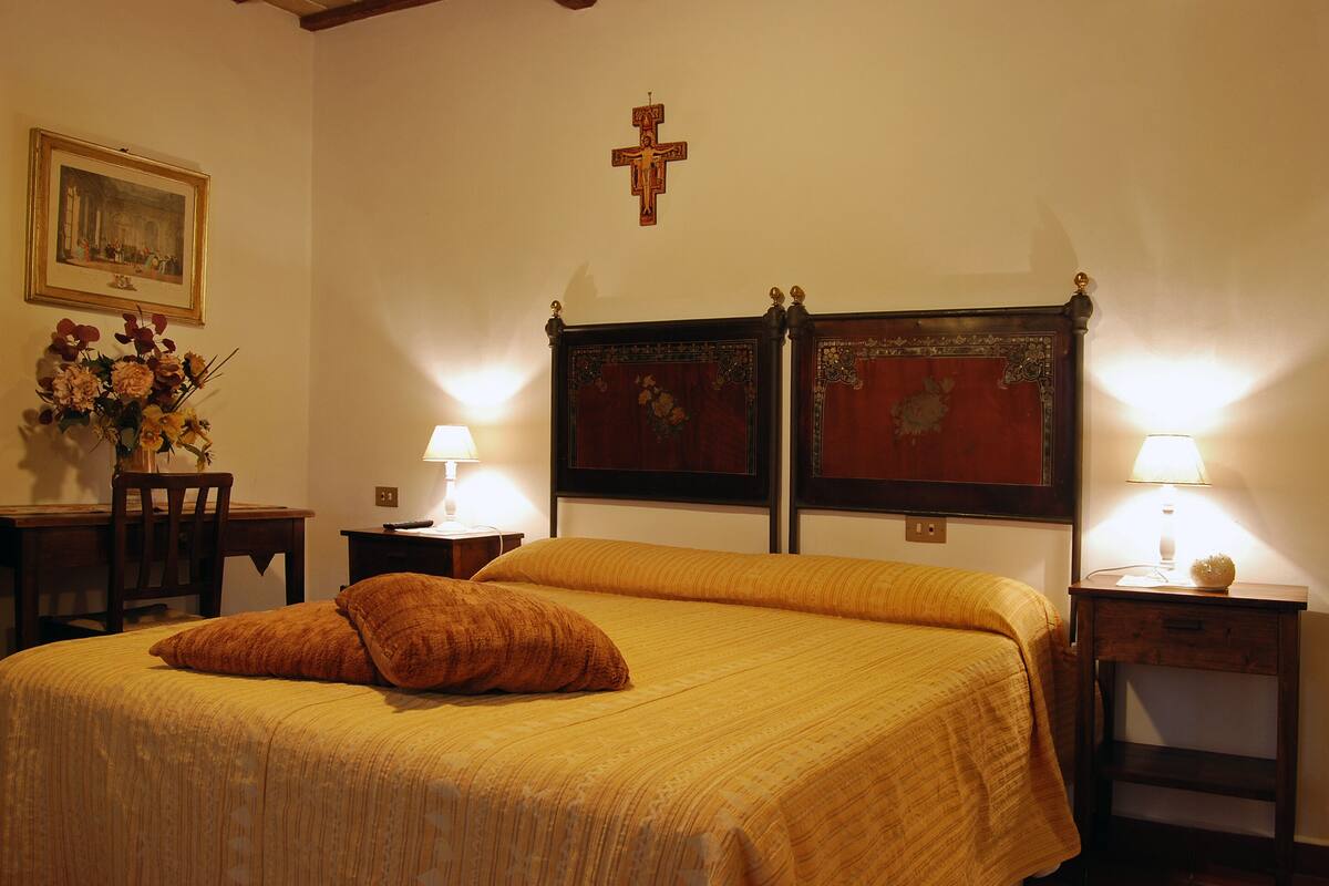 Un lit bien fait, avec une tête de lit, deux lampes de chevet, une croix accrochée au mur et un tableau encadré au-dessus du lit.