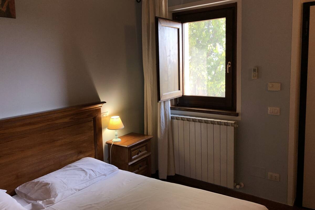Une chambre à coucher avec une tête de lit en bois, un lit avec une literie blanche, une table de chevet avec une lampe, une fenêtre avec des rideaux et un radiateur.
